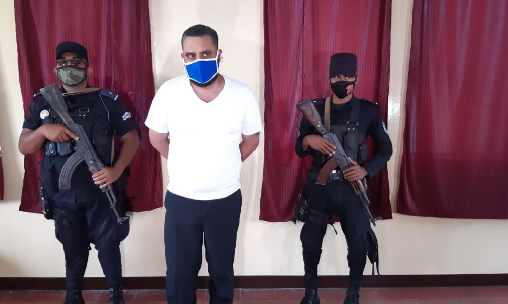 policías con armas y mascarilla custodian a preso con mascarilla azul y camisa blanca